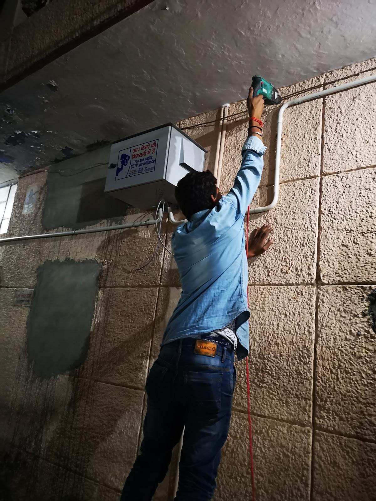 Technician drilling a wall to fix conduit above a CCTV enclosure during a night installation