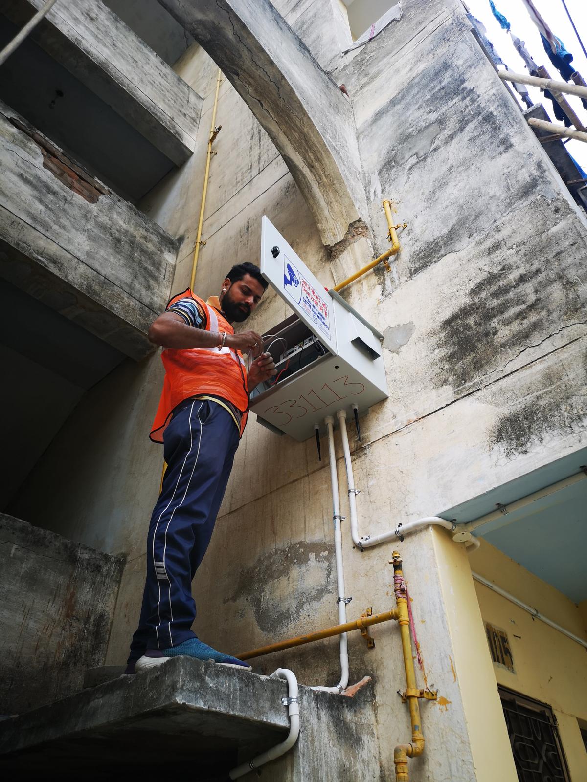 Technician in a safety vest wiring a wall-mounted CCTV equipment enclosure on a building exterior