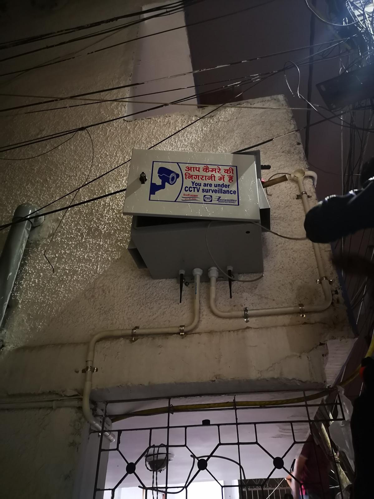 Wall-mounted CCTV equipment enclosure with surveillance signage and conduit routing on a building exterior in Delhi