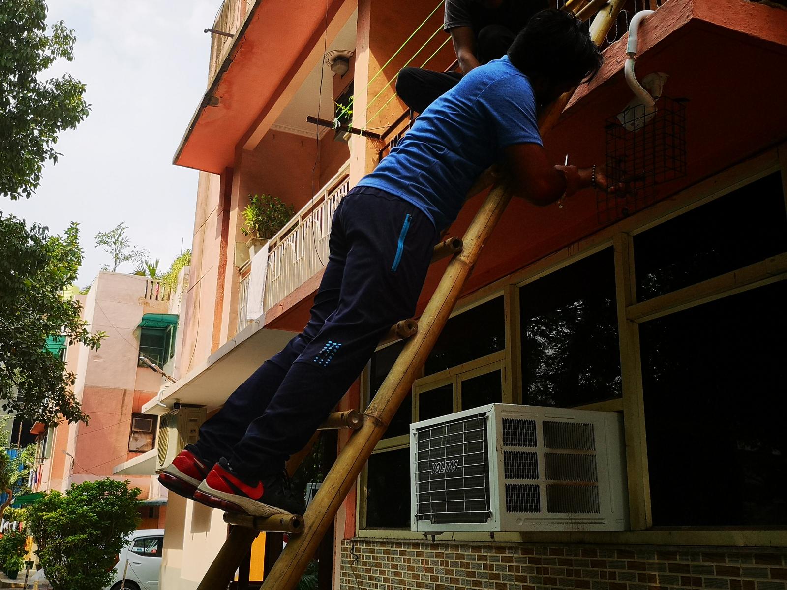 Technician on a bamboo ladder fixing a camera mount on a first-floor wall